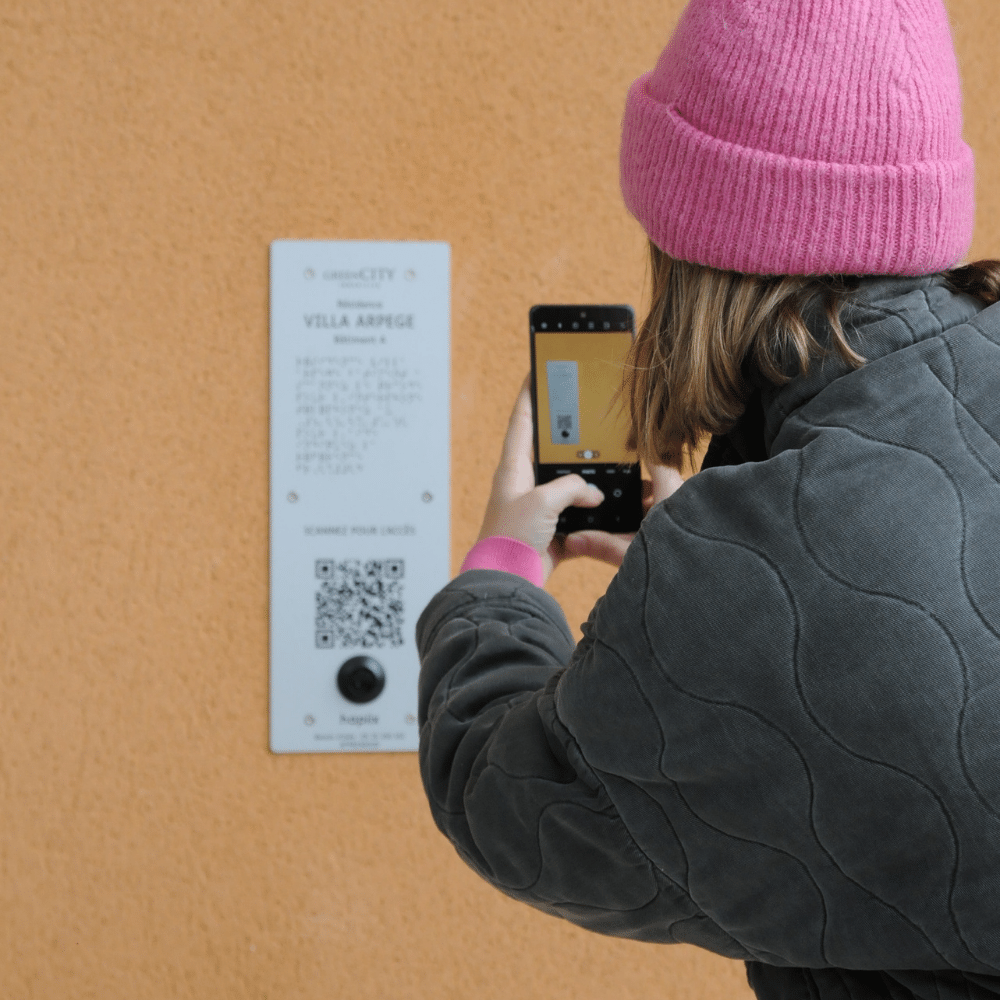 Photo d'une femme en train de scanner avec son smartphone le QR Code gravé sur la plaque hapiix à l'entrée d'un bâtiment.