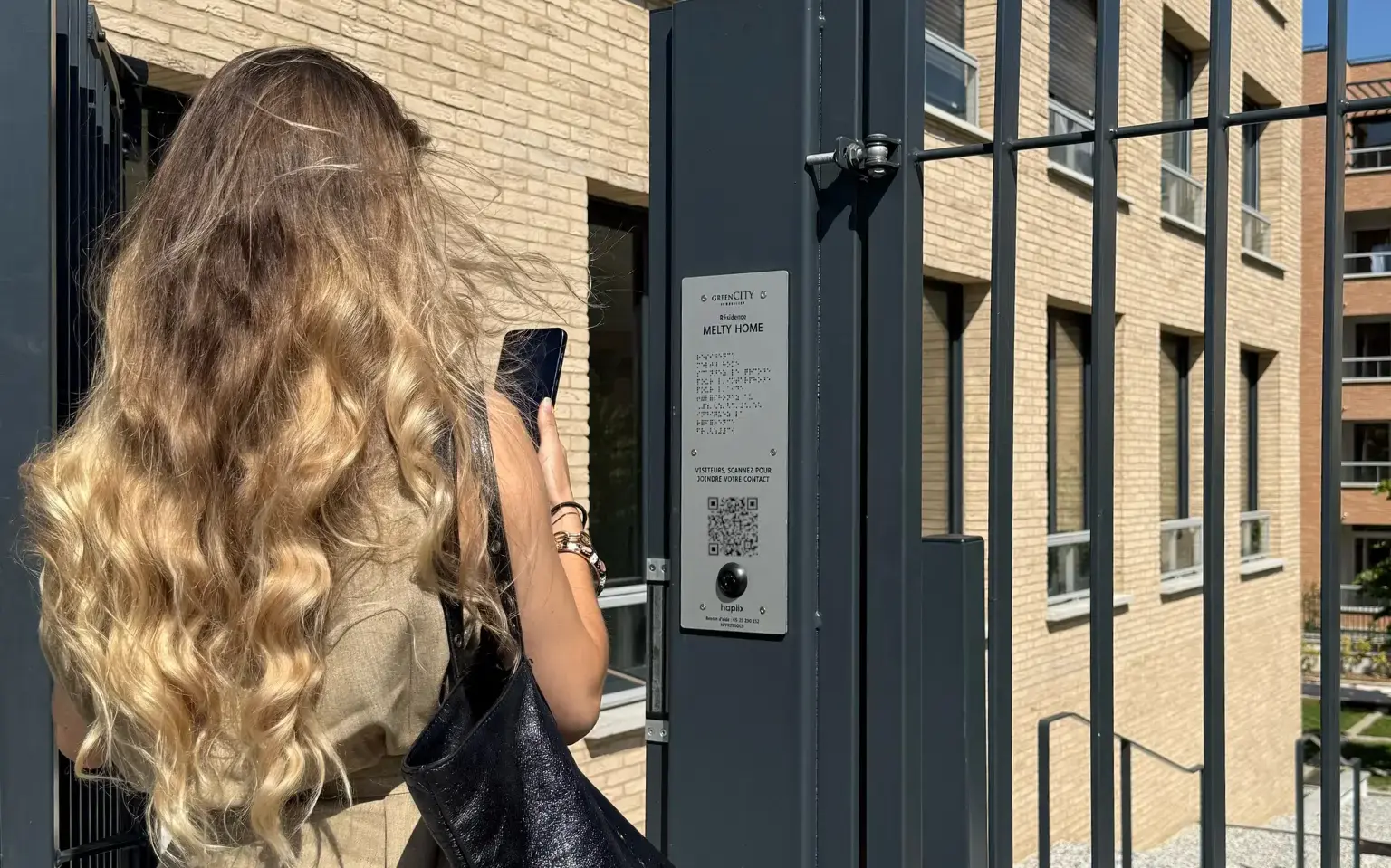 Photo d'une résidente qui rentre chez elle grâce au système hapiix. On la voit le téléphone à la main en train de scanner le QR code  sur la plaque à l'entrée de la résidence. 